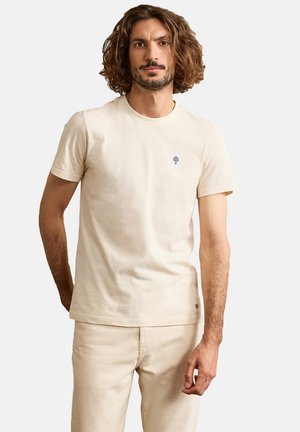 Man with shoulder-length hair wearing a plain beige short-sleeve T-shirt and matching beige pants, standing against a white background.