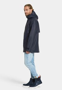 Marinblå vattentät jacka med huva, med en avslappnad passform och kardborreförsedda ärmslut, kombinerad med ljusblå jeans och svarta stövlar som når till ankeln.