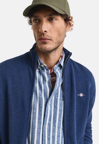 Marineblauer Reißverschluss-Pullover aus strukturiertem Stoff über einem hellblauen und weißen gestreiften Hemd mit Knopfschluss und Brusttasche, das ein Logo zeigt.