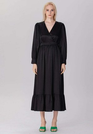 Femme aux longs cheveux blonds portant une robe midi noire à manches longues avec un ourlet à volants et des talons verts à bouts ouverts, debout devant un fond uni.