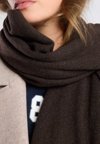Sciarpa a maglia marrone scuro avvolta attorno al collo, con una superficie texture, abbinata a un cappotto beige chiaro con colletto e una camicia blu navy.