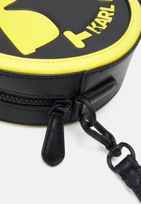 Okrągła czarna skórzana torba z żółtymi akcentami i logo. Posiada zamek błyskawiczny i odpinany czarny pasek. Gładka faktura.