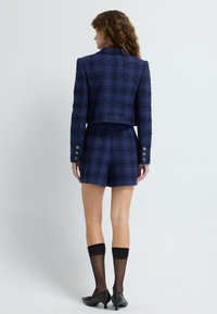 Mörkblå rutigt tweed kort jacka med silverknappar, matchad med shorts i samma tyg. Svarta genomskinliga knästrumpor och låga svarta skor.