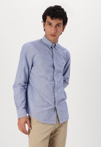 Camisa de botón azul claro con un patrón sutil, mangas largas y un cuello clásico. Combinada con pantalones beige.