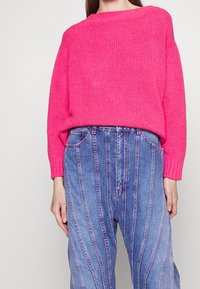 Helles pinkes Strickpullover mit lockerer Passform und gerippten Bündchen, kombiniert mit blauen Jeans aus Denim, die vertikale Streifen und pinke Nähte aufweisen.