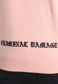 Vaaleanpunainen collegepaita, jossa on ribbaushelma ja joka esittelee rohkeaa mustaa tekstiä "CRIMINAL DAMAGE" alareunassa. Pehmeä, sileä kangas.