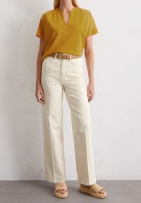Haut jaune moutarde à manches courtes avec un décolleté en V, associé à un pantalon large crème et des sandales tressées tan. Une ceinture simple accentue la taille.