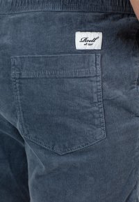 Pantalones de pana azul marino con un bolsillo trasero, que presentan una etiqueta cosida. La textura es acanalada, con sutiles líneas verticales y detalles de costura.