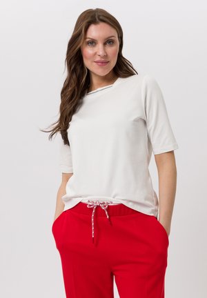 Wit T-shirt met korte mouwen en een ronde hals, voorzien van een decoratieve bies. Gecombineerd met rode broek met een taillekoord.