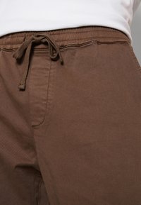 Braune Jogginghose mit Kordelzug und elastischem Bund, mit einer glatten Textur und seitlicher Tasche. Der Stoff wirkt robust und knitterfrei.