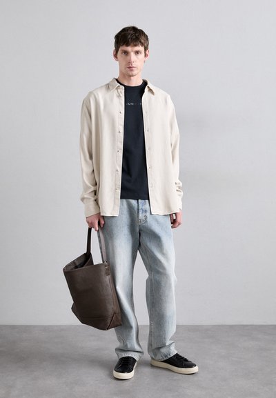 Camisa de botones beige claro sobre una camiseta negra, combinada con jeans sueltos azul claro y zapatillas negras. Se sostiene un bolso de tote de cuero marrón oscuro.