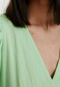 Blouse en tissu vert clair texturé avec un décolleté en V, présentant des plis aux épaules. Pas de motifs distincts ni de garnitures métalliques.