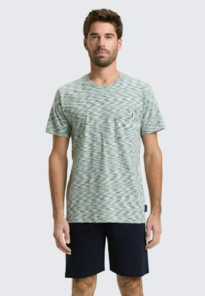 Mężczyzna stojący, ubrany w zielono-biały wzorzysty t-shirt z krótkim rękawem i kieszenią na piersi oraz czarne szorty na jednolitym tle.