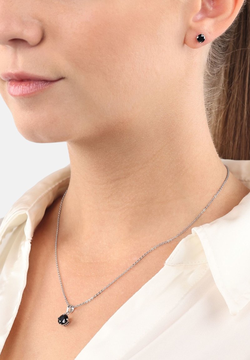 Collana con pendente in cristallo nero e orecchini a bottone abbinati in metallo tono argento; texture liscia; design della catena delicato; stile minimalista.