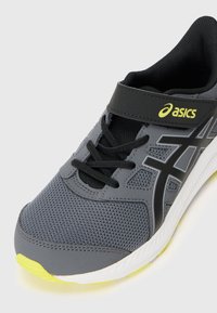 Zapatilla deportiva gris con parte superior de malla, acentos negros y suela amarilla. Tiene cordlaces y una correa de Velcro para un ajuste seguro.