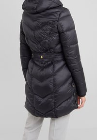 Schwarze Steppjacke mit Chevron-Steppnähten, Kapuze und tailliertem Gürtel. Verfügt über Seitentaschen mit goldfarbenen Metallakzenten. Glatte, glänzende Textur.