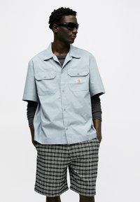 Άνδρας που φοράει μαύρα γυαλιά ηλίου, ανοιχτό μπλε κοντομάνικο πουκάμισο πάνω από μαύρο μακρυμάνικο και ασπρόμαυρο σορτς με καρό σχέδιο.