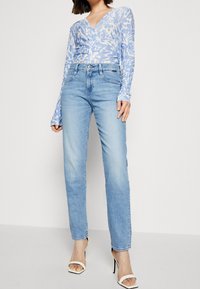 Hellblaues Langarmoberteil mit floralem Muster, kombiniert mit hellblauen gerade geschnittenen Jeans und weißen Absatzsandalen. Glatte Stoffoberfläche.