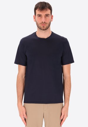 Uomo con capelli corti e scuri che indossa una maglietta blu navy semplice e pantaloni beige, in piedi contro uno sfondo bianco, rivolto in avanti.