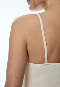 Crème ribbed tanktop met een slanke strap en verstelbare hardware. Soepele textuur met subtiele variaties in toon, die de schouder en arm accentueert.