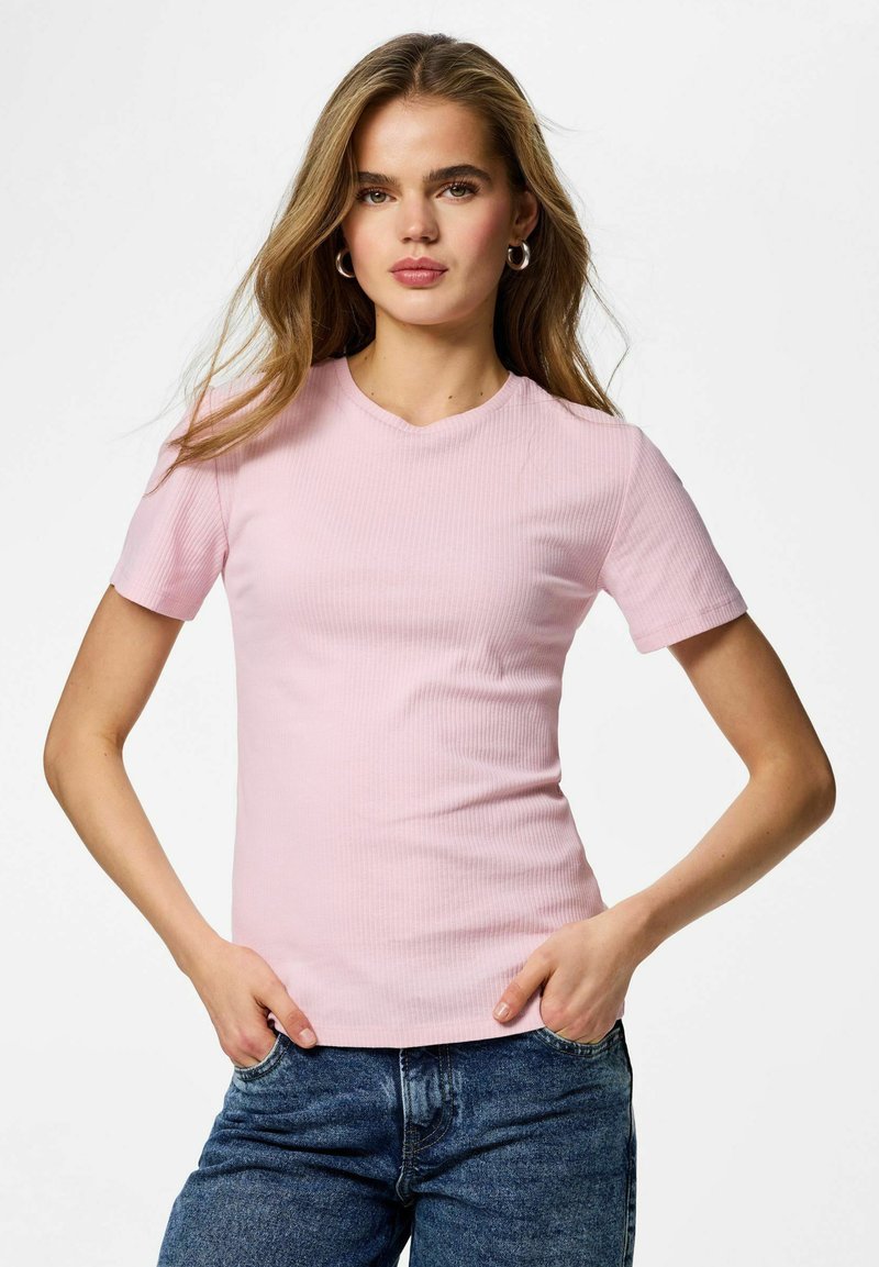 Femme aux cheveux longs portant des boucles d'oreilles en argent en forme d'anneau, un haut côtelé à manches courtes de couleur rose clair, et un jean denim bleu avec les mains dans les poches.