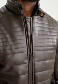 Giacca in pelle marrone con cuciture trapuntate, collo alto, chiusura con zip e dettaglio di bottoni argentati. Texture liscia con design aderente.