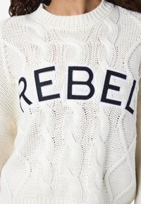 Kreminė kabelių megztinė su juodu siuvinėjimu "REBEL". Turi apvalią kaklą ir tekstūruotą raštą visame. 