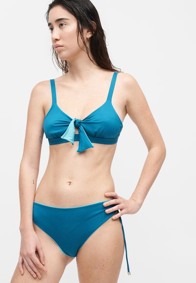 Γυναίκα με νωπά μαλλιά που φορά ένα δίχρωμο μαγιό σε τιρκουάζ με δέσιμο στο μπροστινό μέρος του τοπ, στέκεται μπροστά σε ένα απλό φωτεινό φόντο.