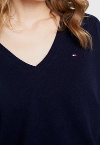 Tmavě modrý svetr s výstřihem do V z měkké látky, s malým červenobíločerveným logem na levé straně hrudi.