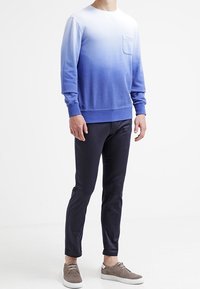 Persona vistiendo un sudadera de manga larga con un degradado de azul a blanco, pantalones oscuros de corte slim y zapatillas de deporte casuales marrón claro, de pie sobre un suelo blanco.