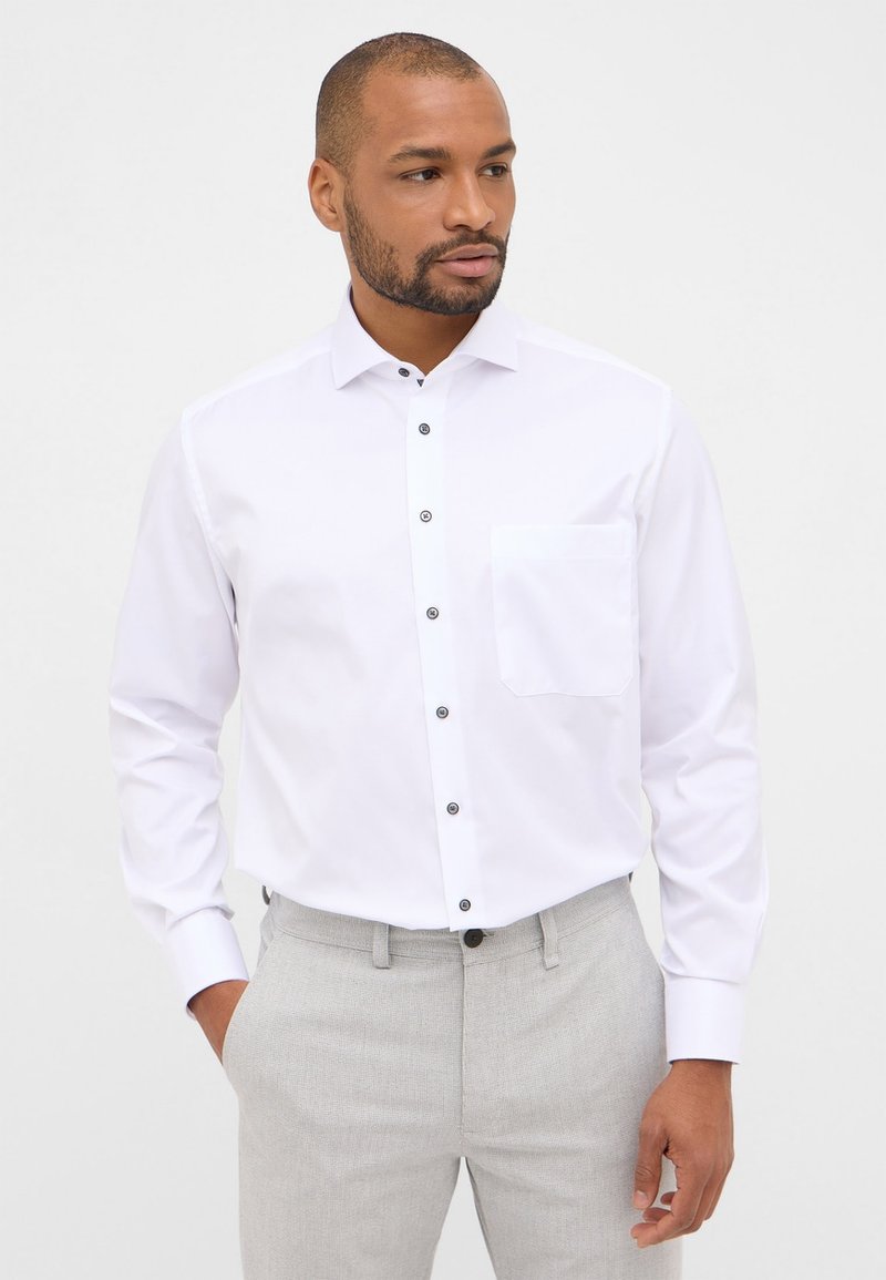 Camicia bianca con bottoni e maniche lunghe, dotata di bottoni neri e taschino sul petto. Realizzata in tessuto morbido, presentata con pantaloni grigi.