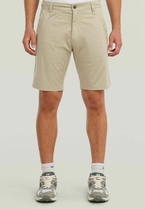 Beige knielange Shorts, getragen von einer stehenden Person, kombiniert mit weißen Socken und grauen Sportschuhen auf neutralem Hintergrund.