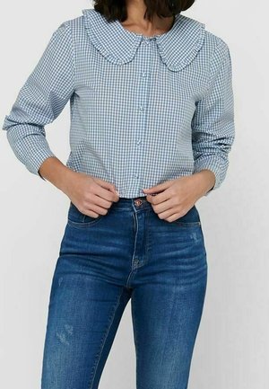 Kobieta w niebiesko-białej bluzce w kratę z dużym falbankowym kołnierzem i niebieskich, wysokich jeansach, ręce poprawiają bluzkę.