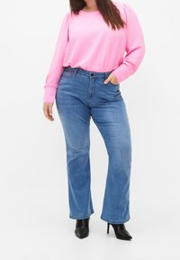 Haut à manches longues rose clair avec tissu texturé, associé à un jean flare bleu en denim taille mi-haute ; des bottes noires à talons hauts complètent le look.