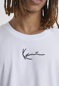 T-shirt en coton blanc avec un design de signature brodé noir sur la poitrine. Col rond et texture douce.