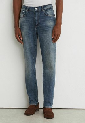 Homme portant un jean bleu délavé droit, une chemise blanche et des mocassins marron, debout contre un mur blanc.