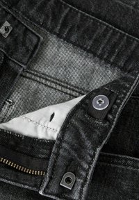 Melnas džinsa džinsu jostasvieta ar regulējamu pogas siksnu, atvērtu rāvējslēdzēju un redzamu baltu auduma kabatu oderi iekšpusē.