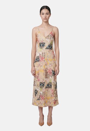 Donna con capelli scuri in un abito midi patchwork floreale con spalline sottili e tacchi beige a punta, in piedi su uno sfondo semplice e chiaro.