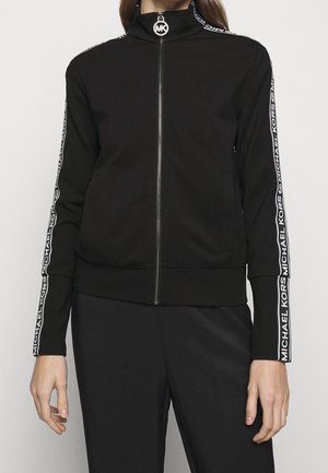 Kobieta nosząca czarną kurtkę zapinaną na zamek błyskawiczny z białym napisem „Michael Kors” na rękawach i okrągłym suwakiem MK, w połączeniu z czarnymi spodniami.