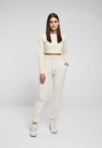 Hellbeige, taillierte Langarmoberteil kombiniert mit hellgrauen Sweatpants, die über einen elastischen Bund und seitliche Taschen verfügen. Lässige weiße Sneakers.