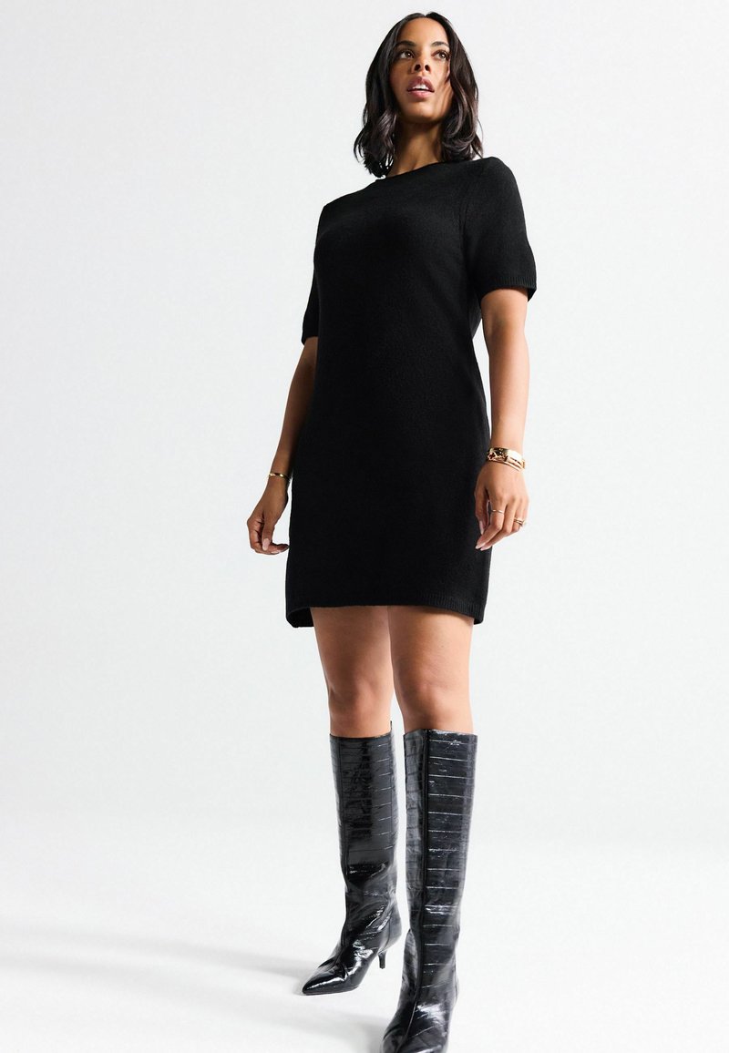 Schwarzes Strickkleid mit kurzen Puffärmeln, einem runden Ausschnitt und einer geraden Silhouette. Kombiniert mit hohen, glänzenden, schwarzen Kniefallstiefeln.