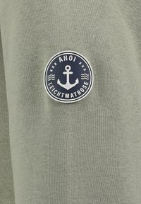 Olijf groene sweatshirtstof met een ronde marineblauwe patch met een ankerontwerp, de tekst "AHOI" en "LEICHTMATROSE," en gedetailleerde stiksels.