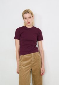 T-shirt bordeaux a maniche corte con scollatura rotonda e un piccolo logo sul petto, abbinato a pantaloni in corduroy beige chiaro a vita alta.