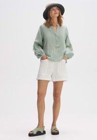 Blusa verde chiaro, strutturata, con scollatura a V; abbinata a pantaloni corti bianchi con tasche frontali e risvolti. Cappello nero e di paglia, sandali verdi.