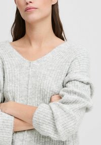 Grauer gerippter Pullover mit V-Ausschnitt, aus weichem, strukturiertem Stoff und voluminösen, umgeschlagenen Ärmeln. Das Design betont Komfort und Schlichtheit.