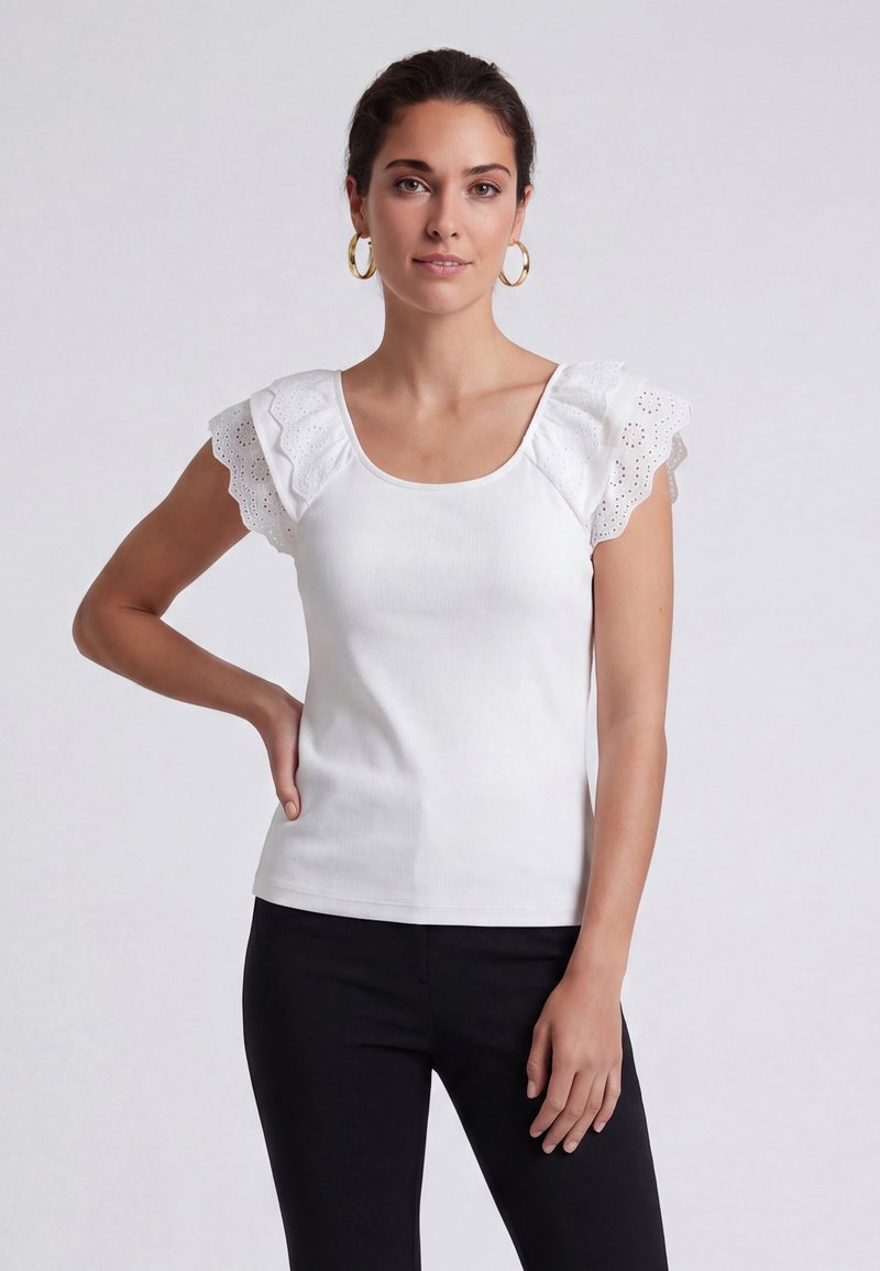 Femme aux cheveux foncés portant un haut blanc avec des manches à volants ajourées et un pantalon noir, debout avec une main sur la hanche sur un fond uni.
