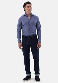 Camicia a maniche lunghe con fantasia blu e chiusura a bottoni, abbinata a jeans in denim scuro con risvolti, indossata con scarpe nere.