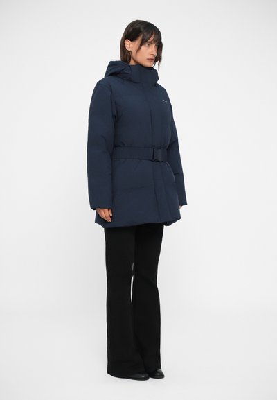 Manteau rembourré bleu marine avec une taille ceinturée, une capuche et des manches longues. Présente une texture lisse et un design minimaliste. Associé à un pantalon noir évasé.