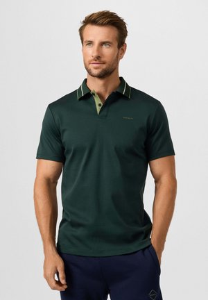 Man draagt een donkergroene polo met korte mouwen en contrasterende kraagrand en donkerblauwe broek, staand tegen een effen lichte achtergrond.