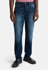 Granatowe dżinsy o prostym kroju, z efektem utlenienia i pięcioma kieszeniami, w połączeniu z czarnymi skórzanymi butami.